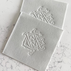 May include: Two square, white fabric labels with a raised, ornate monogram design. The monogram features intertwined letters and decorative flourishes. The labels have a textured surface and are stacked slightly, showcasing the design.