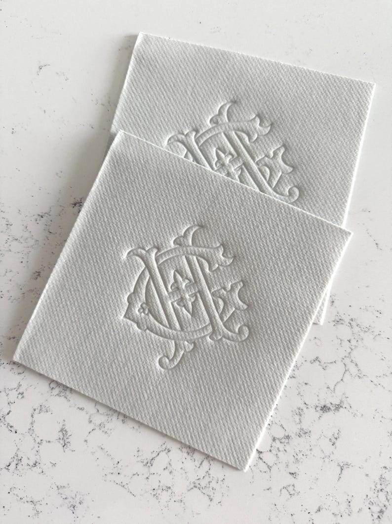May include: Two square, white fabric labels with a raised, ornate monogram design. The monogram features intertwined letters and decorative flourishes. The labels have a textured surface and are stacked slightly, showcasing the design.