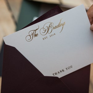 May include: A white thank you card with gold lettering that reads "The Bradleys EST 2019" and "THANK YOU". The card is in a burgundy envelope.