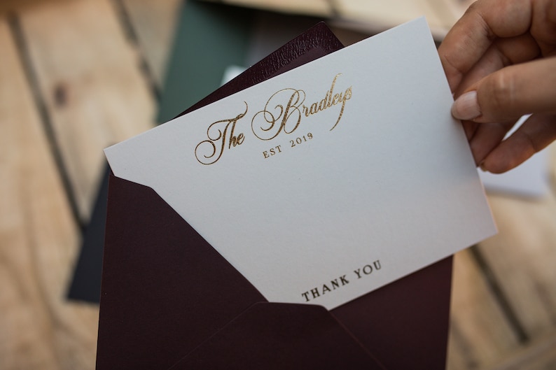 May include: A white thank you card with gold lettering that reads "The Bradleys EST 2019" and "THANK YOU". The card is in a burgundy envelope.