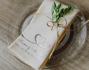 May include: A white linen napkin with a silver-colored design of two intertwined hearts and the text "Kathrin & Matthias" and "Schon dass du mit uns feierst". The napkin is tied with twine and decorated with a sprig of green leaves.