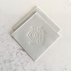 May include: Two white square coasters with a textured surface. Each coaster features an embossed monogram design in the center. The coasters are arranged in a diamond shape, with one slightly overlapping the other. The background is a white marble surface.