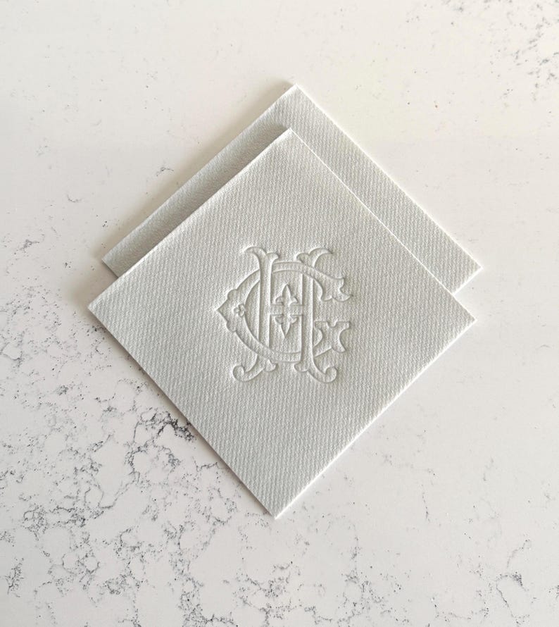 May include: Two white square coasters with a textured surface. Each coaster features an embossed monogram design in the center. The coasters are arranged in a diamond shape, with one slightly overlapping the other. The background is a white marble surface.