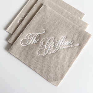 Peut inclure: Une pile de trois serviettes carr&eacute;es beiges avec un texte brod&eacute; en blanc indiquant The Griffins et la date du 27 septembre 2025. Les serviettes pr&eacute;sentent une texture de tissu tiss&eacute; visible et sont dispos&eacute;es en quinconce.