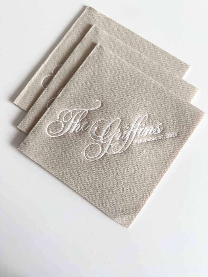 Peut inclure: Une pile de trois serviettes carr&eacute;es beiges avec un texte brod&eacute; en blanc indiquant The Griffins et la date du 27 septembre 2025. Les serviettes pr&eacute;sentent une texture de tissu tiss&eacute; visible et sont dispos&eacute;es en quinconce.