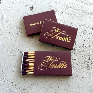 May include: Burgundy matchboxes with gold lettering. One box reads "March 11," and others display "The Smiths" in elegant script. One box is open, revealing matches with purple tips.