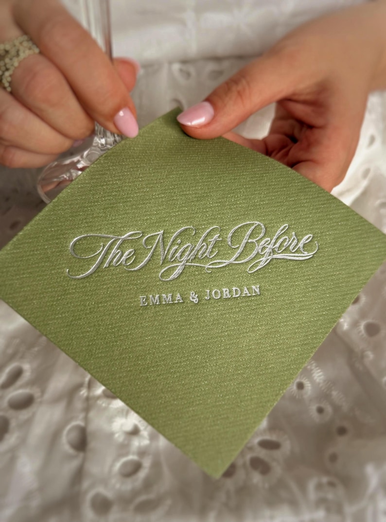 Op de afbeelding: Een vierkant, saliegroen papieren servet met een textuur, voorzien van de zilveren reli&euml;ftekst The Night Before en de namen EMMA & JORDAN in een kleiner, strak schreefloos lettertype dat eronder is gedrukt.