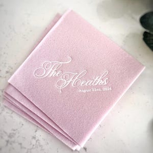 Peut inclure: Une pile de serviettes en papier carr&eacute;es rose clair avec du texte imprim&eacute; en blanc. Le texte affiche les mots The Heaths dans une grande police cursive, avec la date du 22 ao&ucirc;t 2026 imprim&eacute;e dans une police sans empattement plus petite.