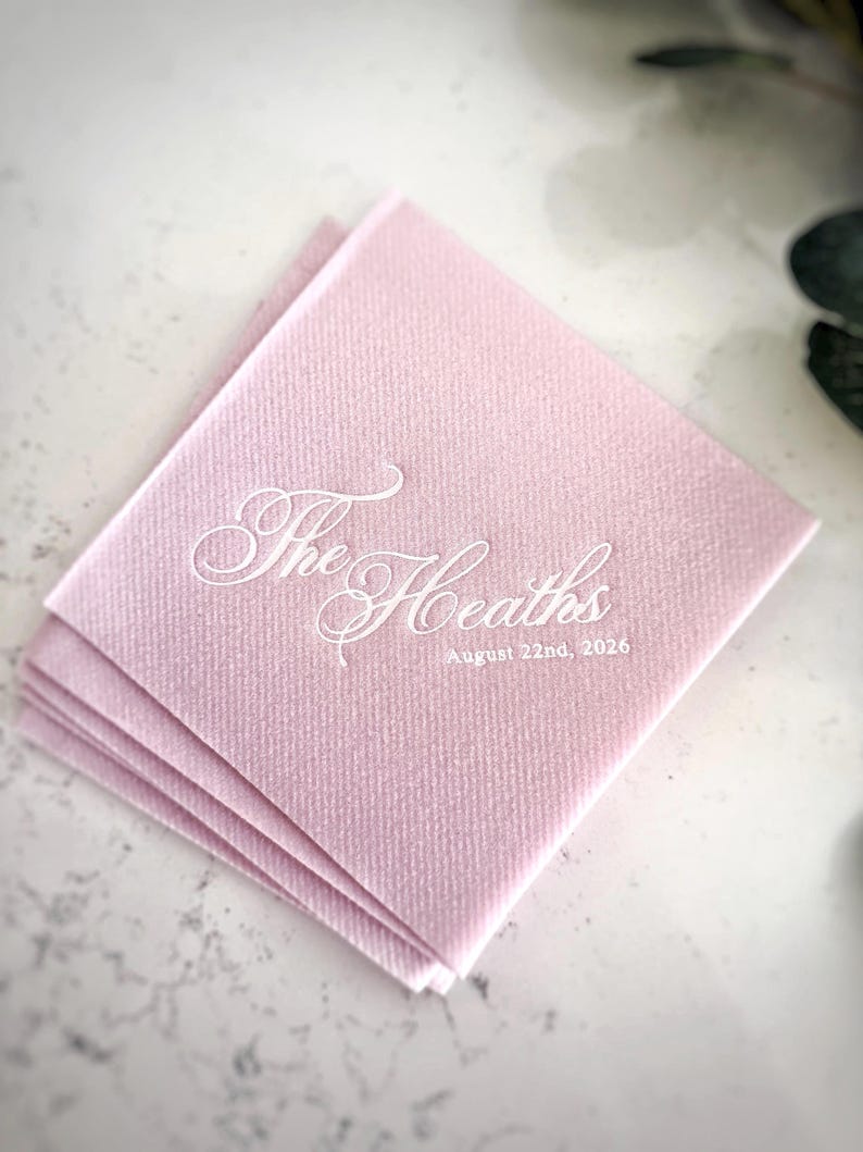 Peut inclure: Une pile de serviettes en papier carr&eacute;es rose clair avec du texte imprim&eacute; en blanc. Le texte affiche les mots The Heaths dans une grande police cursive, avec la date du 22 ao&ucirc;t 2026 imprim&eacute;e dans une police sans empattement plus petite.