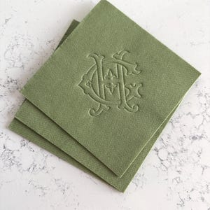May include: Two olive green cloth napkins stacked on a white surface. Each napkin features an embossed monogram design in a matching olive green color. The napkins have a square shape and a textured appearance.