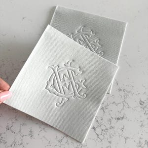 May include: Two white square coasters with an embossed monogram design. The coasters are made of a textured material and are arranged on a light-colored surface. The monogram appears to be a stylized combination of letters.