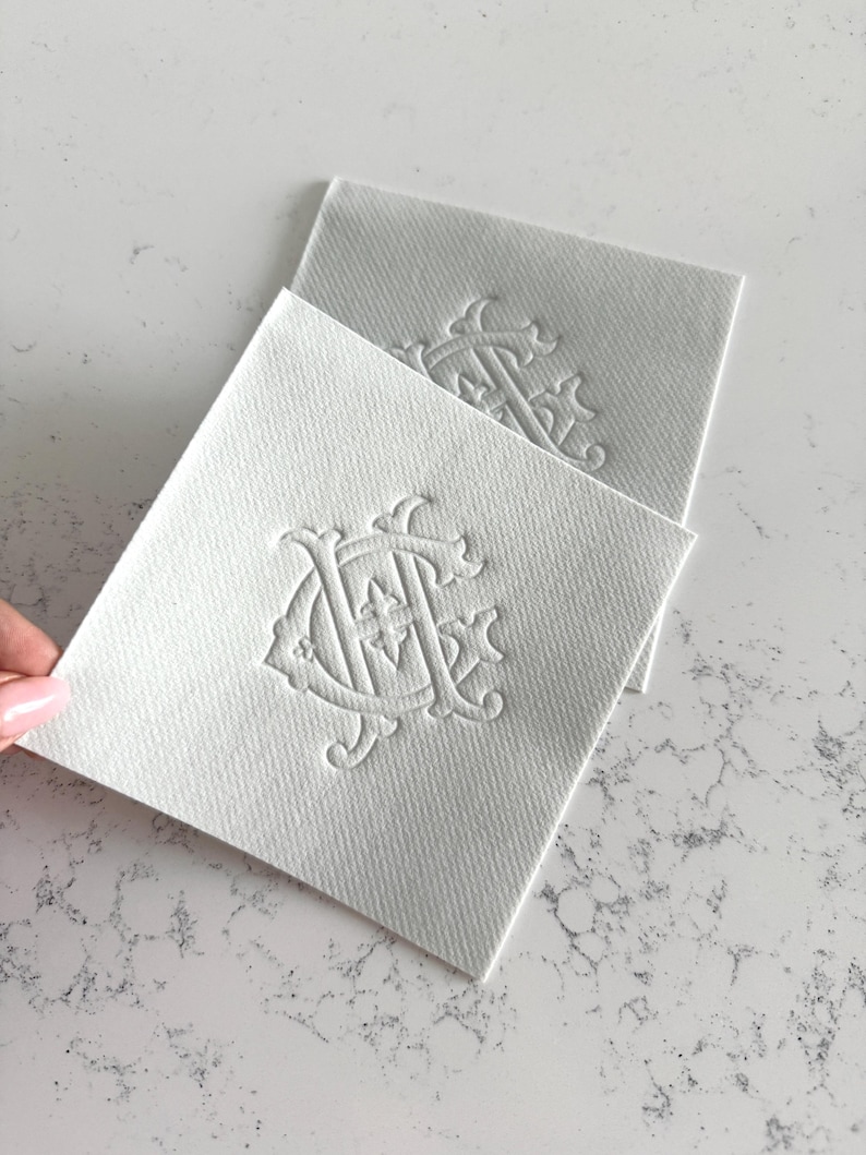 May include: Two white square coasters with an embossed monogram design. The coasters are made of a textured material and are arranged on a light-colored surface. The monogram appears to be a stylized combination of letters.