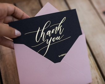 May include: A black card with gold lettering that says "thank you". The card is in a pink envelope.