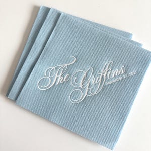 Peut inclure: Une pile de serviettes en papier carr&eacute;es bleu clair avec un texte blanc en relief indiquant The Griffins et September 27, 2025. Les serviettes pr&eacute;sentent une surface textur&eacute;e et sont dispos&eacute;es en pile l&eacute;g&egrave;rement d&eacute;cal&eacute;e et superpos&eacute;e.