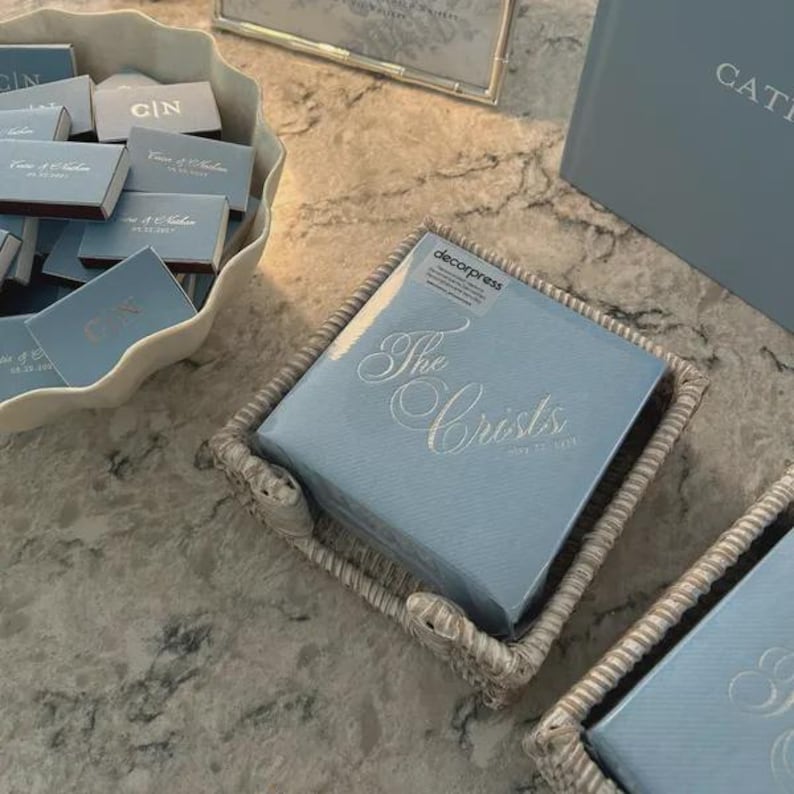Peut inclure: Une pile de serviettes en papier bleu clair avec le texte dor&eacute; The Crists May 22 2027 dans un panier tress&eacute;, &agrave; c&ocirc;t&eacute; d'un bol festonn&eacute; rempli de petites bo&icirc;tes d'allumettes bleu clair avec du texte dor&eacute; et les initiales C|N.