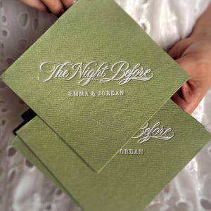 Op de afbeelding: Een vierkante olijfgroene kaart met een textuur die op stof lijkt. De tekst "The Night Before" is in wit schrift in reli&euml;f aangebracht, gevolgd door de namen "EMMA & JORDAN" in een kleiner, strak wit lettertype. Een tweede kaart is zichtbaar.