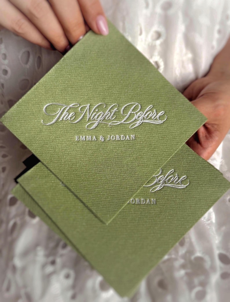 Op de afbeelding: Een vierkante olijfgroene kaart met een textuur die op stof lijkt. De tekst "The Night Before" is in wit schrift in reli&euml;f aangebracht, gevolgd door de namen "EMMA & JORDAN" in een kleiner, strak wit lettertype. Een tweede kaart is zichtbaar.