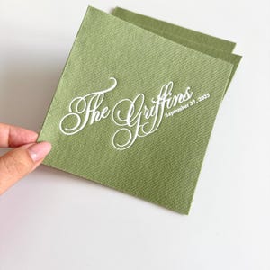 Peut inclure: Une serviette en papier carr&eacute;e de couleur vert sauge avec une surface textur&eacute;e. La serviette pr&eacute;sente un texte imprim&eacute; en blanc dans une &eacute;criture cursive indiquant The Griffins, avec la date 27 septembre 2025 en dessous en police sans empattement.