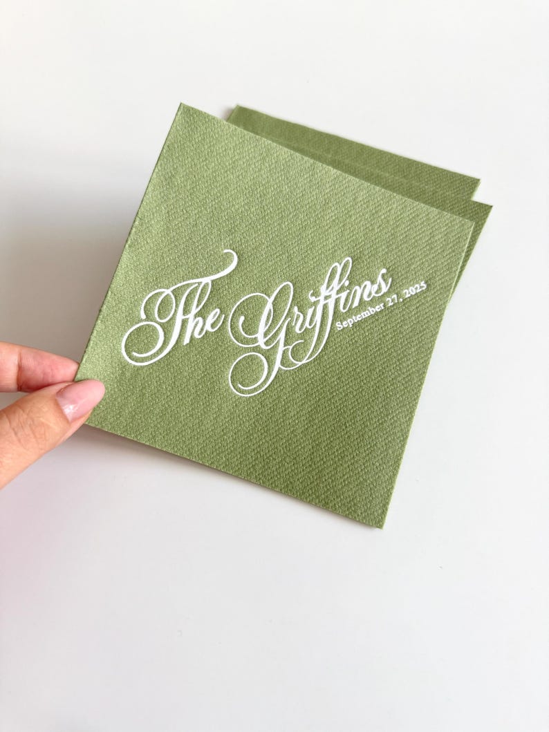 Peut inclure: Une serviette en papier carr&eacute;e de couleur vert sauge avec une surface textur&eacute;e. La serviette pr&eacute;sente un texte imprim&eacute; en blanc dans une &eacute;criture cursive indiquant The Griffins, avec la date 27 septembre 2025 en dessous en police sans empattement.