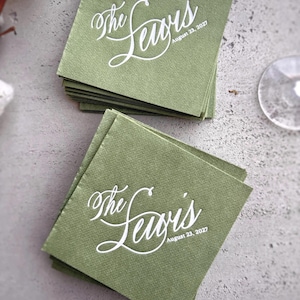 Peut inclure: Plusieurs serviettes carrées vert olive empilées, chacune imprimée avec le texte blanc "The Lewis" et la date "August 23, 2027". Les serviettes sont posées sur une surface grise. L'arrière-plan est flou.