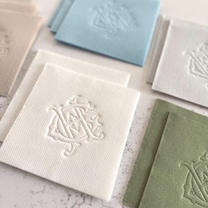May include: A collection of square fabric coasters in various colors, including white, light blue, and green. Each coaster features an embossed monogram design. The coasters are arranged on a light-colored surface.