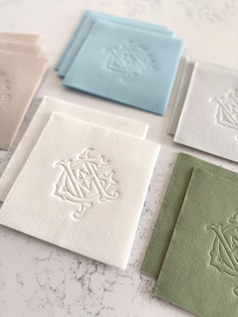 May include: A collection of square fabric coasters in various colors, including white, light blue, and green. Each coaster features an embossed monogram design. The coasters are arranged on a light-colored surface.