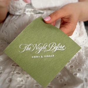 Op de afbeelding: Een vierkant saliegroen papieren servet met witte reli&euml;ftekst die The Night Before leest in een cursief lettertype, gevolgd door de namen EMMA & JORDAN in een strak, hoofdletter schreefloos lettertype onder de hoofdtitel.