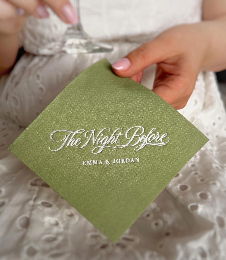 Op de afbeelding: Een vierkant saliegroen papieren servet met witte reli&euml;ftekst die The Night Before leest in een cursief lettertype, gevolgd door de namen EMMA & JORDAN in een strak, hoofdletter schreefloos lettertype onder de hoofdtitel.