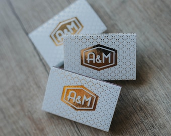 May include: Three rectangular white matchboxes featuring a repeating gold geometric pattern and a central gold hexagonal emblem with the letters A&M printed in white. The boxes are arranged in a cluster, showcasing the metallic foil finish.