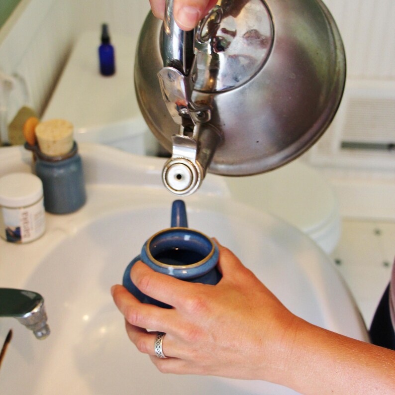 May include: A silver kettle pouring hot water into a blue ceramic teapot. The teapot is sitting in a white porcelain sink.