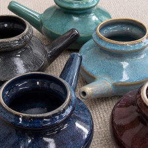 May include: A collection of five ceramic teapots with side handles, featuring glossy glazes in shades of teal, charcoal gray, light blue, deep navy blue, and dark brown. Each teapot has a wide circular opening and a short, tapered spout.