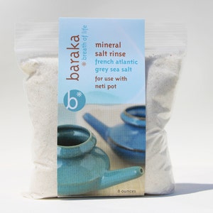 May include: A white plastic bag filled with grey sea salt labelled "baraka * breath of life *" and "mineral salt rinse french atlantic grey sea salt for use with neti pot". The bag is sitting on a white surface. The label also includes a blue and white illustration of a neti pot. The bag contains 8 ounces of salt.