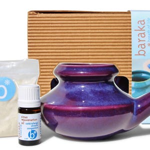 Puede incluir: Un kit de cuidado sinusal Baraka que incluye un neti pot de cerámica morada, un frasco pequeño de vidrio ámbar con aceite sinusal y tapa blanca, y una bolsa de plástico transparente con sal marina mineral, dentro de una caja de cartón.