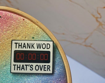 May include: A round embroidery hoop with a rainbow glitter background. A rectangular pin with the text "THANK WOD 00:00:00 THAT'S OVER" is attached to the hoop. The hoop has a wooden frame.