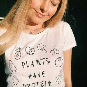 May include: White t-shirt featuring a black line drawing of various fruits and vegetables, such as pineapple, avocado, broccoli, cherries, watermelon, pear, tomato, and bananas. The text "PLANTS HAVE PROTEIN" is printed below.