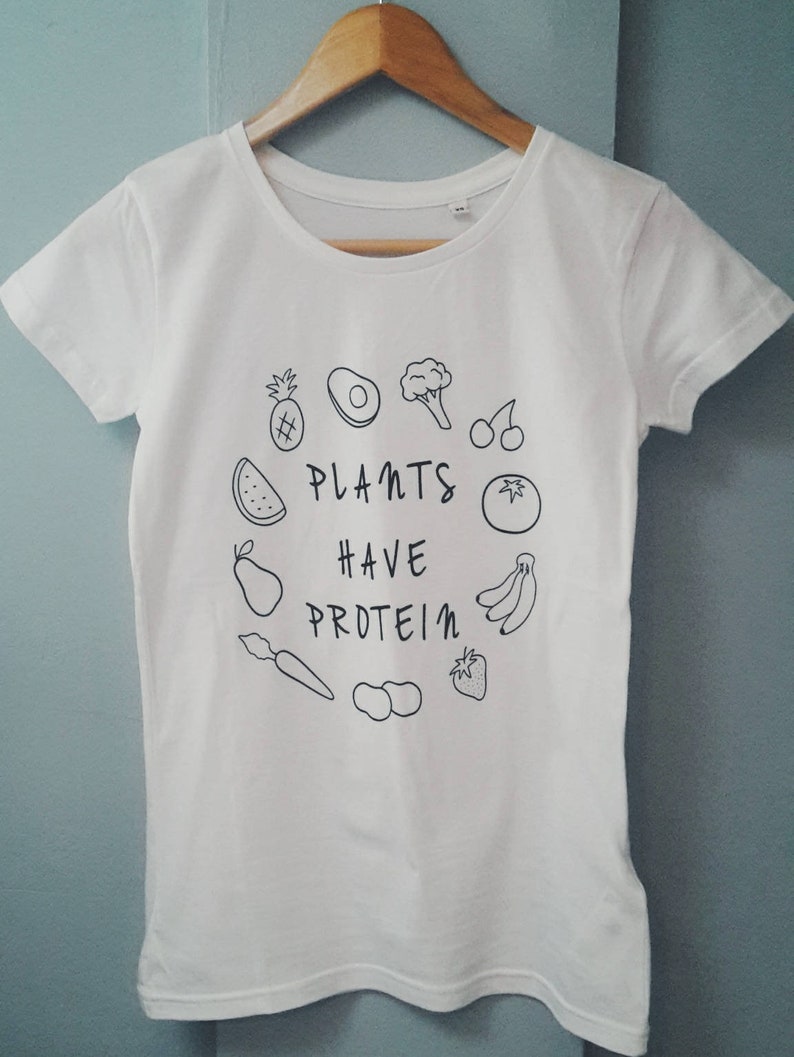 May include: White t-shirt with a round neck and short sleeves. The shirt displays a black line drawing of fruits and vegetables, including a pineapple, avocado, and banana, with the text "PLANTS HAVE PROTEIN".