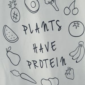 May include: White t-shirt featuring a black outline design of fruits and vegetables, such as a pineapple, avocado, broccoli, cherries, tomato, bananas, and a strawberry. The words "PLANTS HAVE PROTEIN" are written in black.