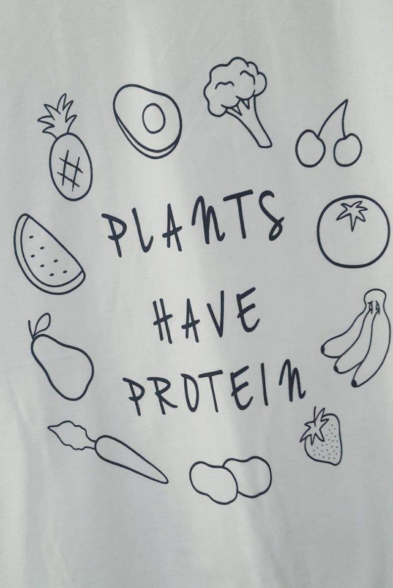 May include: White t-shirt featuring a black outline design of fruits and vegetables, such as a pineapple, avocado, broccoli, cherries, tomato, bananas, and a strawberry. The words "PLANTS HAVE PROTEIN" are written in black.