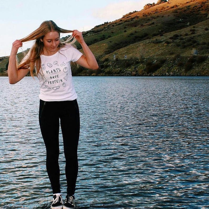 May include: A white t-shirt with the text "PLANTS HAVE PROTEIN" and plant illustrations, worn with black leggings and black and white trainers. The person is standing by a lake, with a mountain in the background.