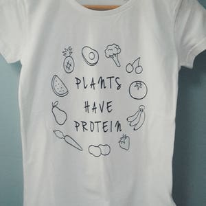 May include: White t-shirt with a round neck and short sleeves. The shirt displays a black line drawing of fruits and vegetables encircling the text "PLANTS HAVE PROTEIN". The design includes a pineapple, avocado, broccoli, cherries, watermelon, pear, carrot, peanuts, strawberry, tomato, and bananas.