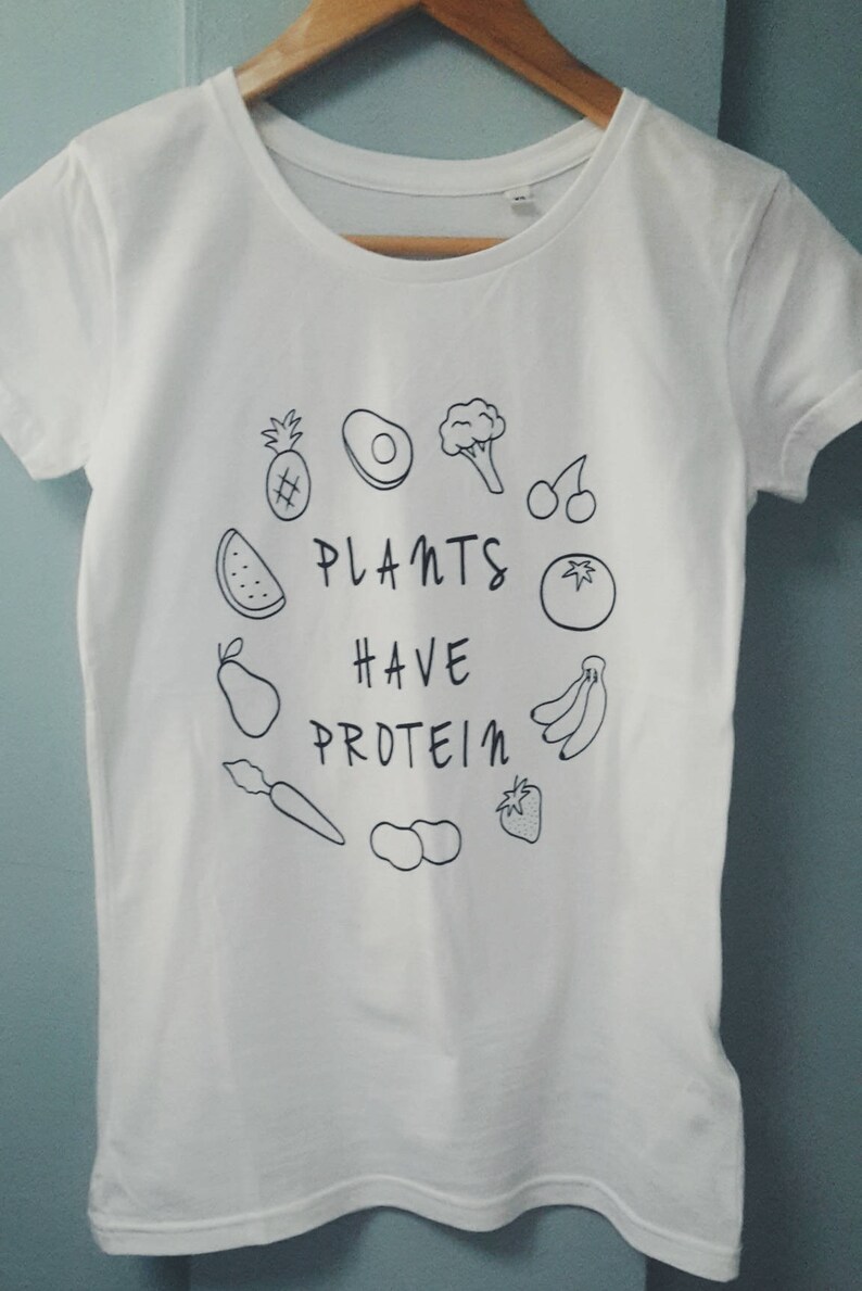 May include: White t-shirt with a round neck and short sleeves. The shirt displays a black line drawing of fruits and vegetables encircling the text "PLANTS HAVE PROTEIN". The design includes a pineapple, avocado, broccoli, cherries, watermelon, pear, carrot, peanuts, strawberry, tomato, and bananas.