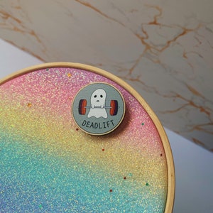 Può includere: Spilla rotonda con un fantasma dei cartoni animati che solleva pesi, la parola "DEADLIFT" è sotto. La spilla è su uno sfondo glitterato arcobaleno all'interno di un telaio da ricamo in legno.