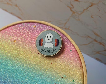 May include: A round pin with a cartoon ghost lifting weights, the word "DEADLIFT" is below the ghost. The pin is on a rainbow glitter background inside a wooden embroidery hoop.