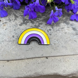 Könnte beinhalten: Ein Ansteck-Pin mit der Non-Binary-Pride-Flagge in Regenbogen-Design. Der Pin hat ein gelbes, weißes, lila und schwarzes Farbschema. Der Pin befindet sich auf einer grauen Betonoberfläche mit blauen Blumen im Hintergrund.