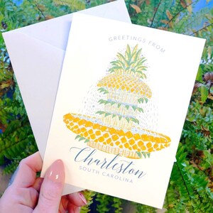 May include: A cream-coloured greeting card featuring a pineapple illustration and the words "Greetings From Charleston South Carolina." The pineapple is yellow and green, with a detailed design. The card is accompanied by a white envelope.