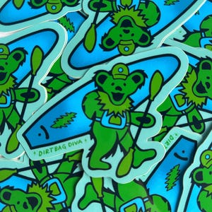 May include: A stack of colorful stickers featuring a cartoon bear character paddling a blue kayak. The stickers have the text "Dirtbag Diva +" on them.