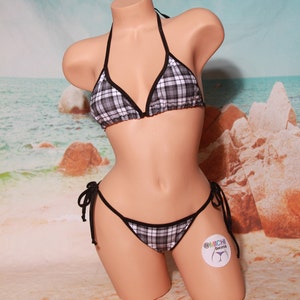 Puede incluir: Conjunto de bikini de dos piezas con estampado de cuadros blancos y negros. La parte superior del bikini es de estilo halter con ribetes negros, y la parte inferior es de estilo tanga. El bikini se exhibe en un maniquí con un fondo de escena de playa.
