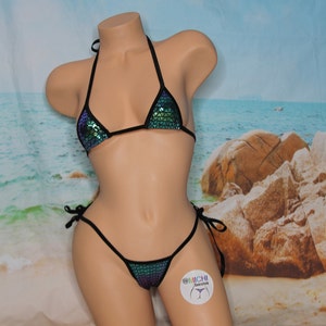 May include: A black bikini with a green and blue iridescent sequin fabric. The bikini top is a triangle style and the bottom is a cheeky style with ties on the sides. The bikini is displayed on a mannequin against a beach background.
