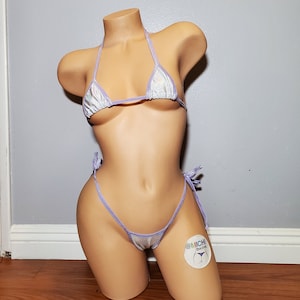 Może przedstawiać: Połyskujący srebrny i lawendowy zestaw bikini. Trójkątny top i dół z wiązaniem są związane lawendowym sznurkiem. Bikini jest prezentowane na manekinie. Ogólnym motywem jest odzież kąpielowa.