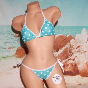 Könnte beinhalten: Ein türkisfarbener Bikini mit weißen Punkten und weißen Rändern. Das Bikinioberteil ist im Dreieckstil gehalten, das Unterteil ist ein freches Modell mit Bändern an den Seiten. Der Bikini befindet sich auf einer Schaufensterpuppe vor einem Strandhintergrund.
